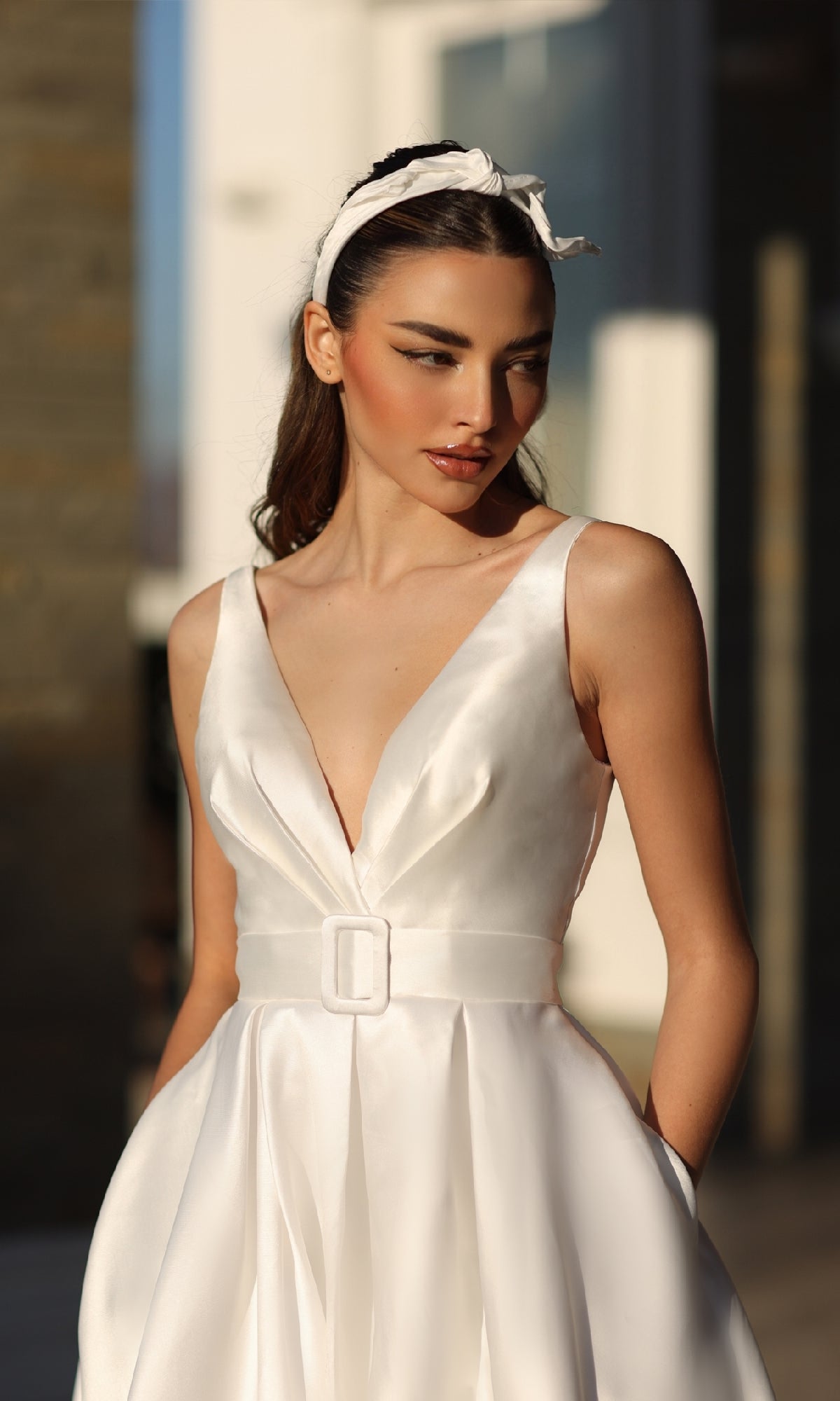 A woman stands outdoors in sunlight wearing the Tina Holly BB209W, a sleeveless long white prom dress with a deep V-neckline, belted waist, and white headband.